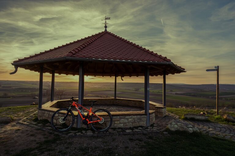 Pavillon mit rotem Dach, Fahrrad davor, Landschaft und Sonnenuntergang im Hintergrund.