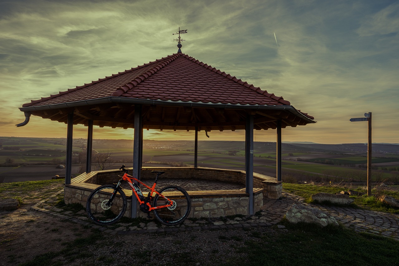 Pavillon mit rotem Dach, Fahrrad davor, Landschaft und Sonnenuntergang im Hintergrund.